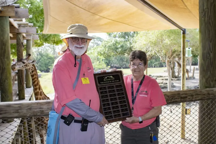 Two older adults stand holding a plaque together. 