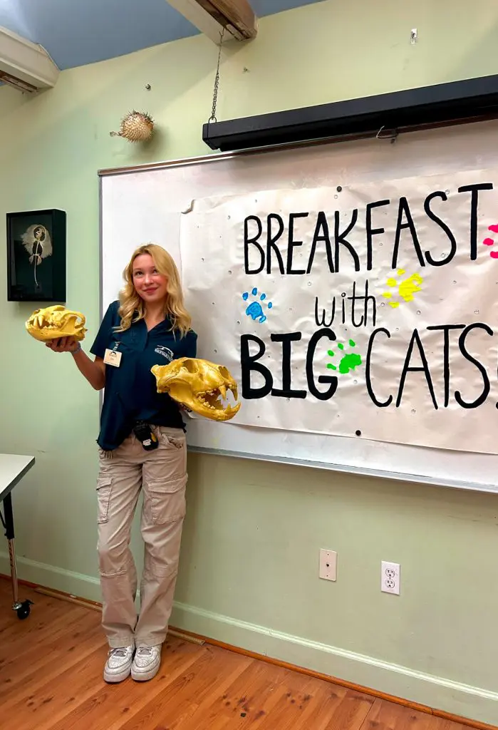 A girl poses with animal anatomy skulls next to a whiteboard that reads "breakfast with the big cats."