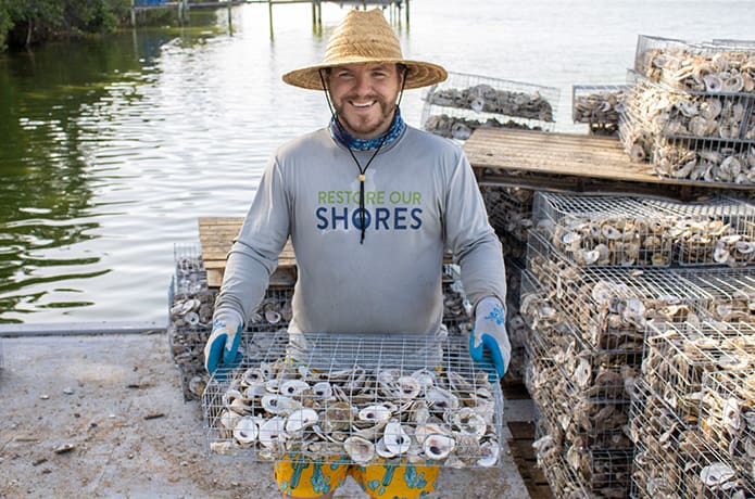 A man holds a wire basket filled with oysters in front of the river.