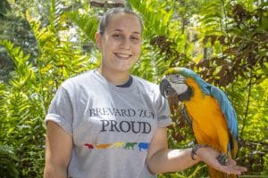 A woman in a Brevard Zoo Proud shirt poses with a macaw. 