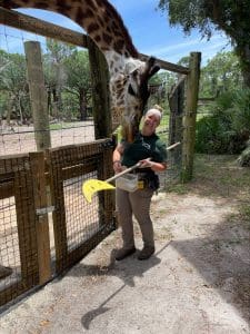 Keeper Makayla stands near a fence with a giraffe nosing her side.