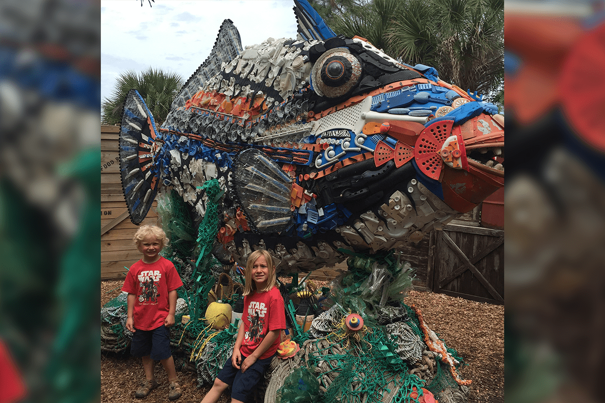 two kids posing in front of a washed ashore artwork fish created from trash recovered from our waterways