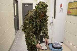 A man wear s a ghillie suit while picking up two saucers of milk.